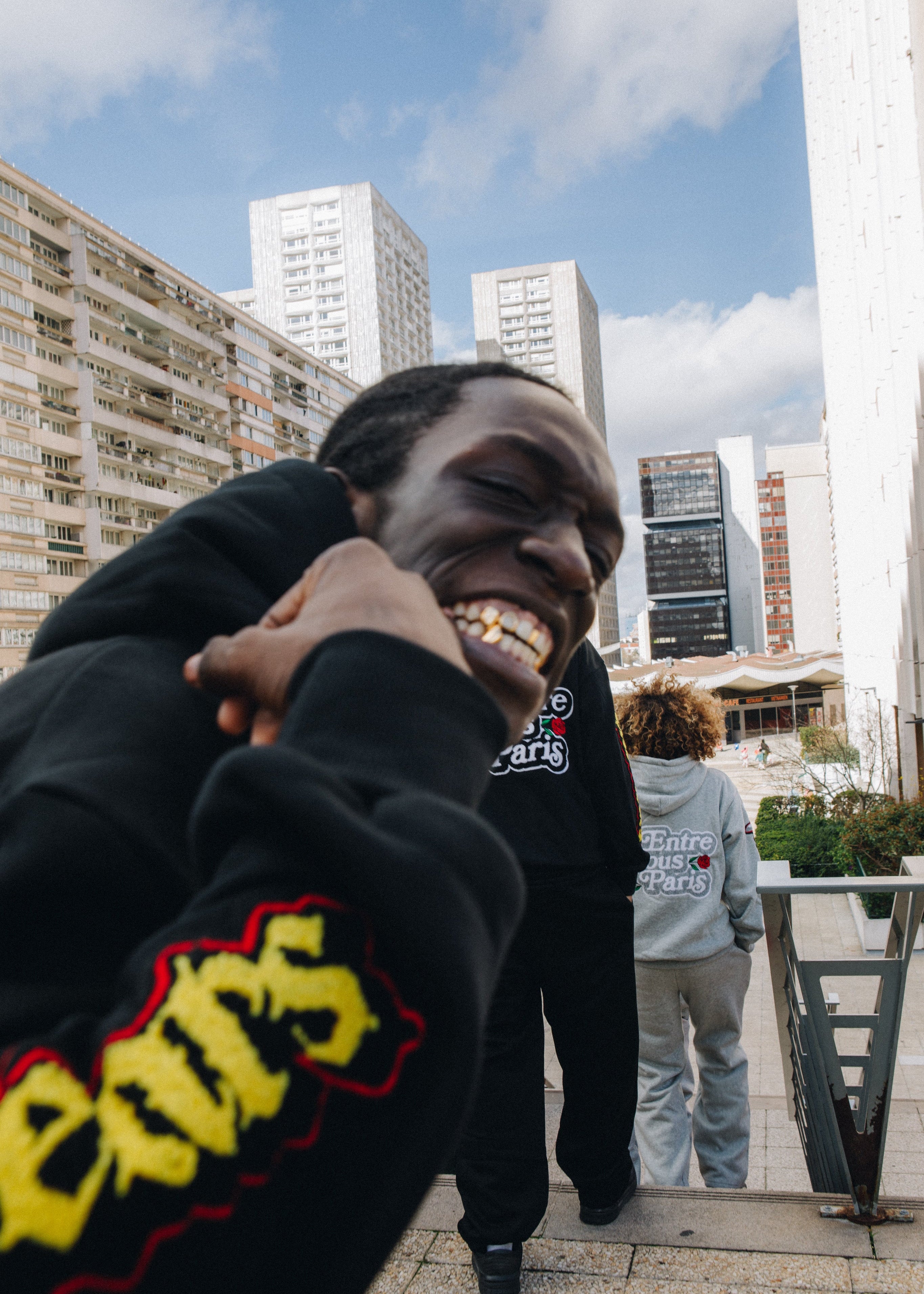 Person in black hoodie with yellow text outdoors with buildings in the background