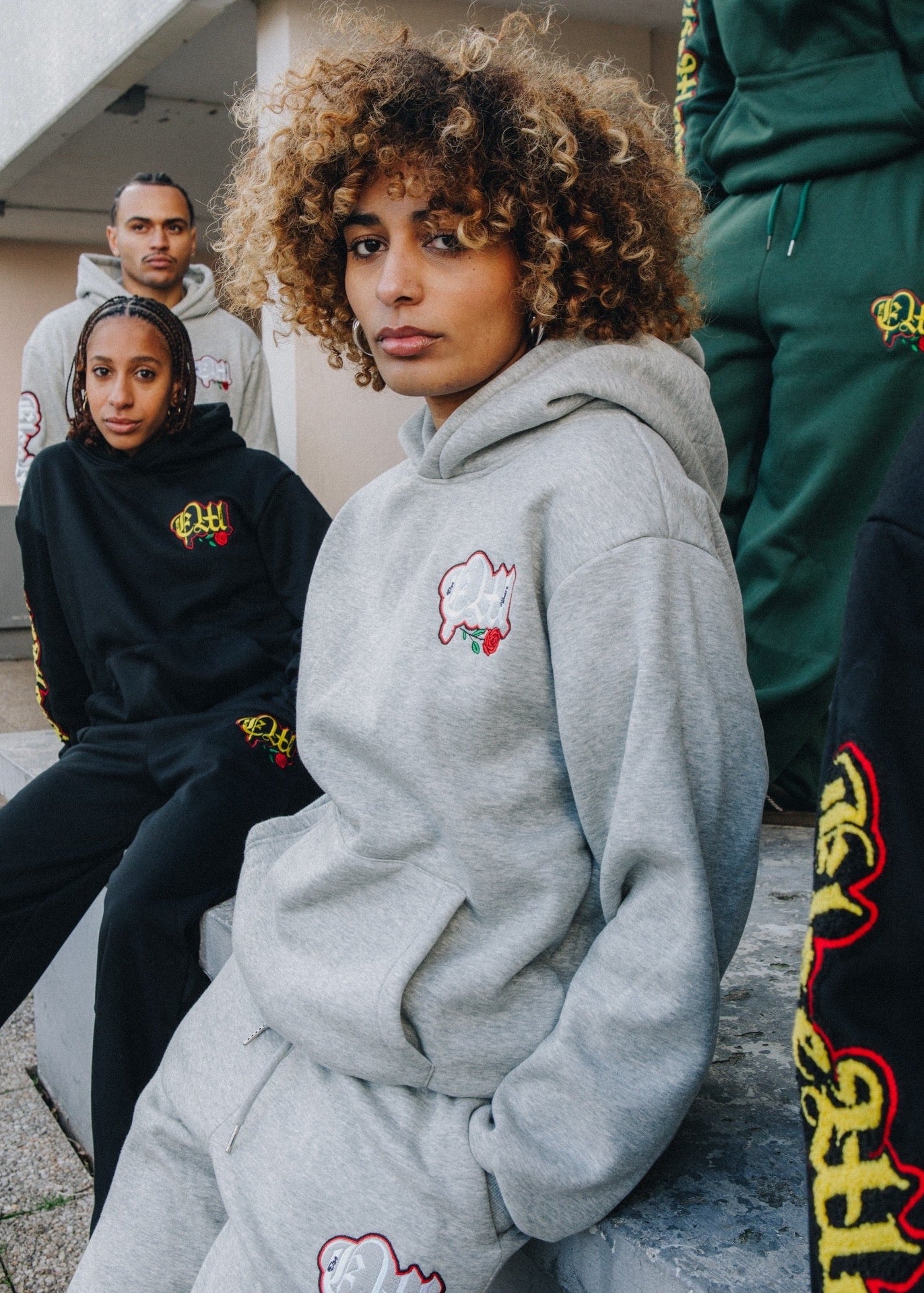 Person wearing a gray hoodie with a logo, sitting outdoors with others in the background.