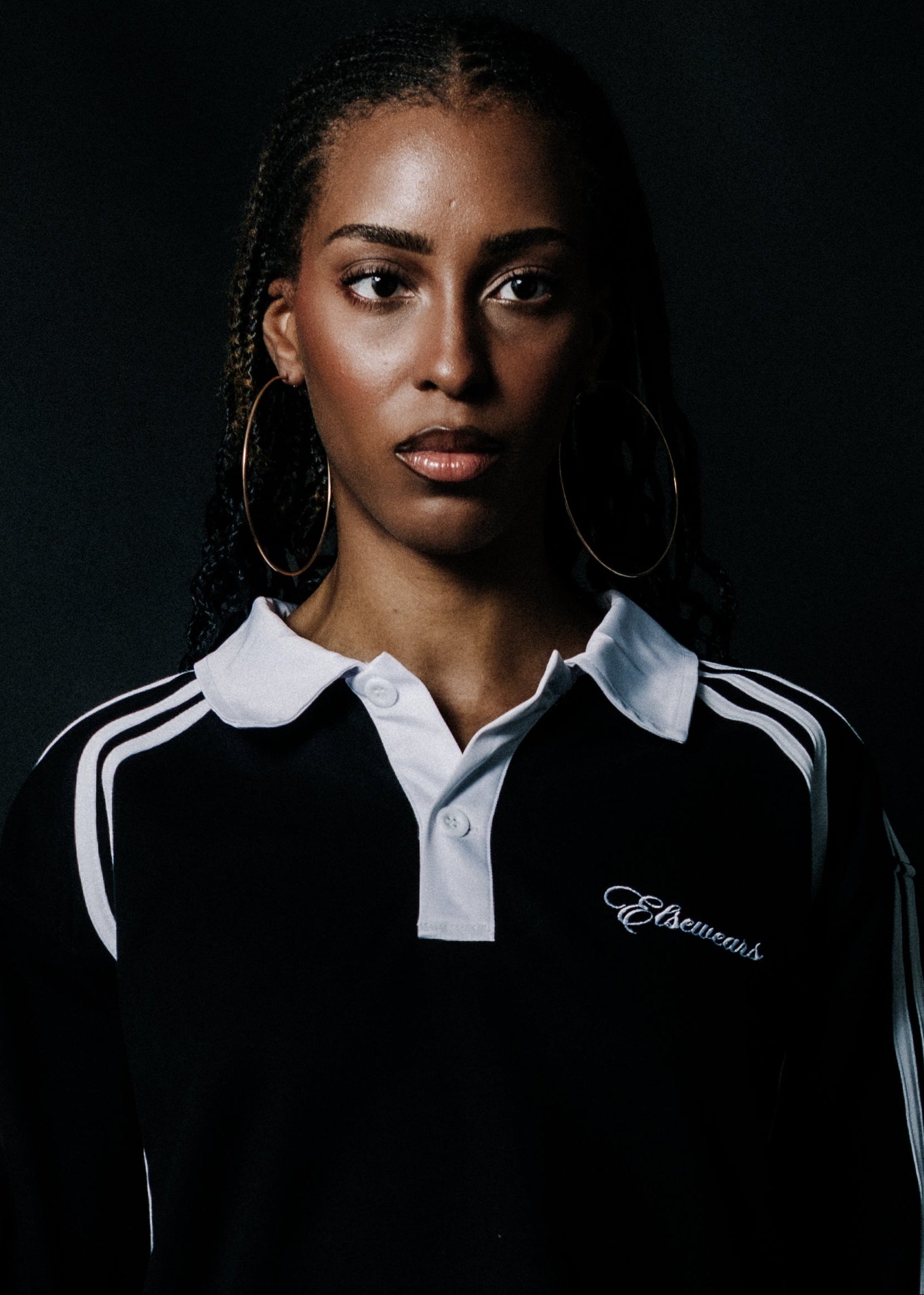 A woman with braided hair wearing a black Adidas track jacket with white stripes and a white collar, set against a dark background.