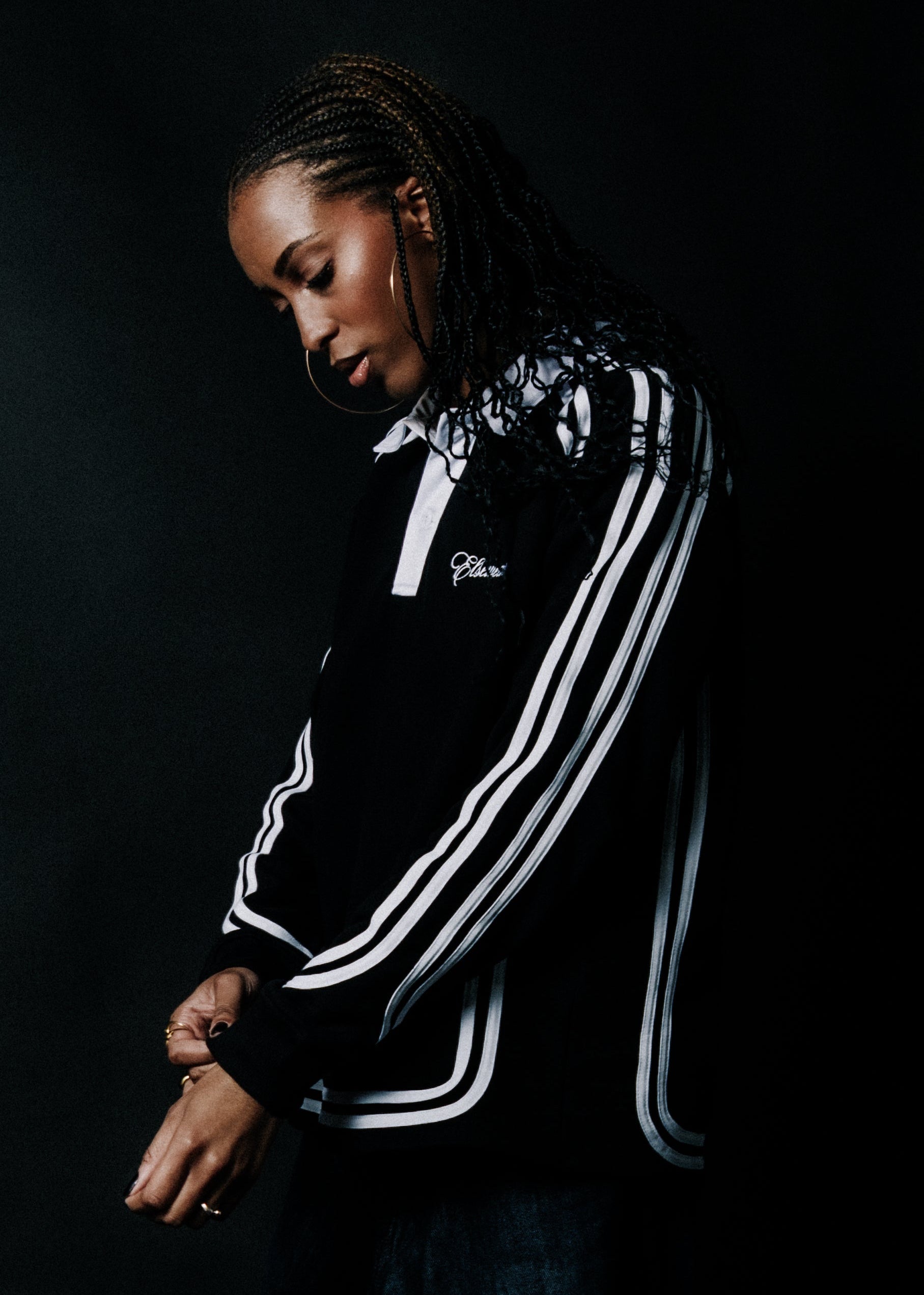 A woman wearing a black Adidas track jacket with white stripes, posed against a dark studio background.